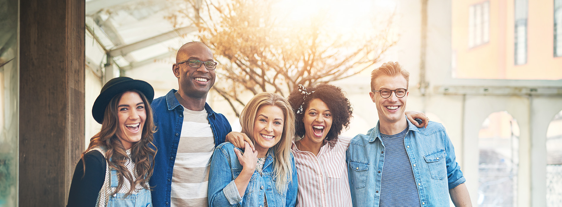 A group of five young adults posing together outdoors with smiles on their faces, standing close together with a cityscape in the background.
