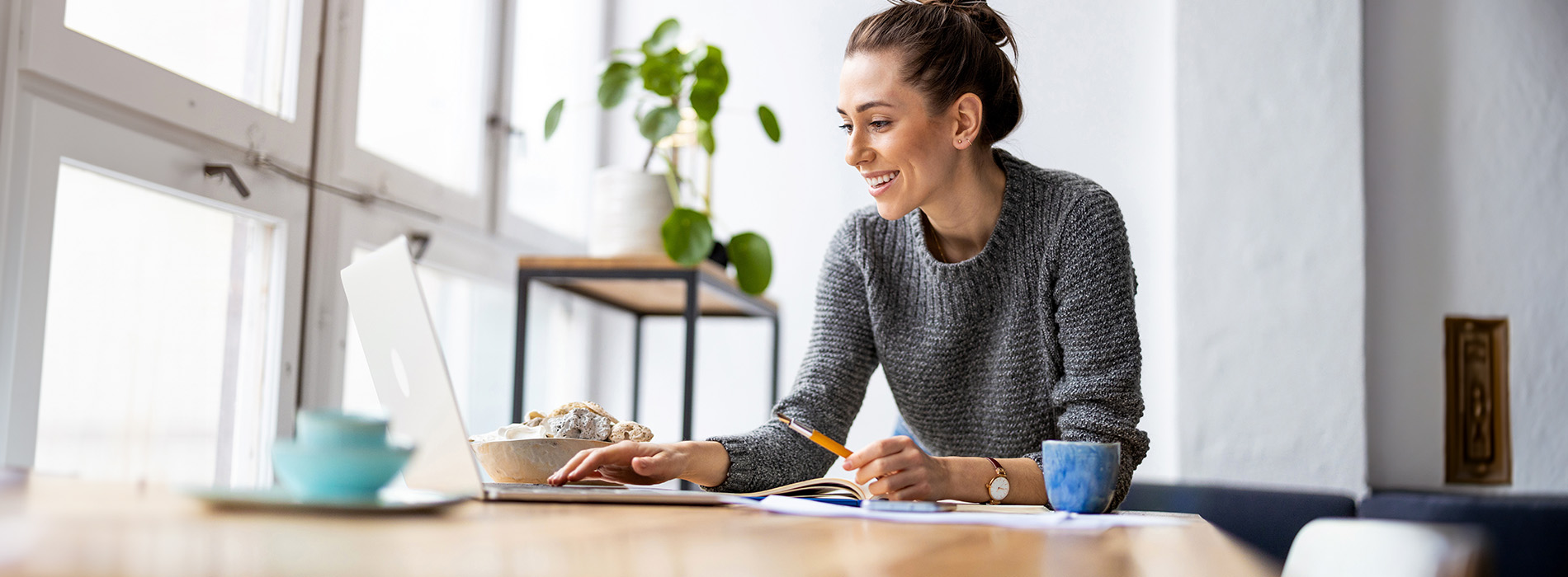 A woman sitting at a desk with a laptop, writing on paper, in a well-lit room.
