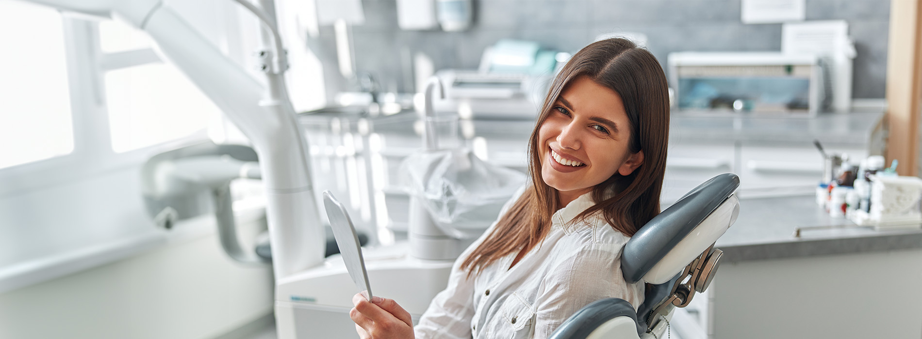 A woman sitting in a dental chair with a smile, holding a clipboard and pen, wearing a white lab coat, surrounded by dental equipment.