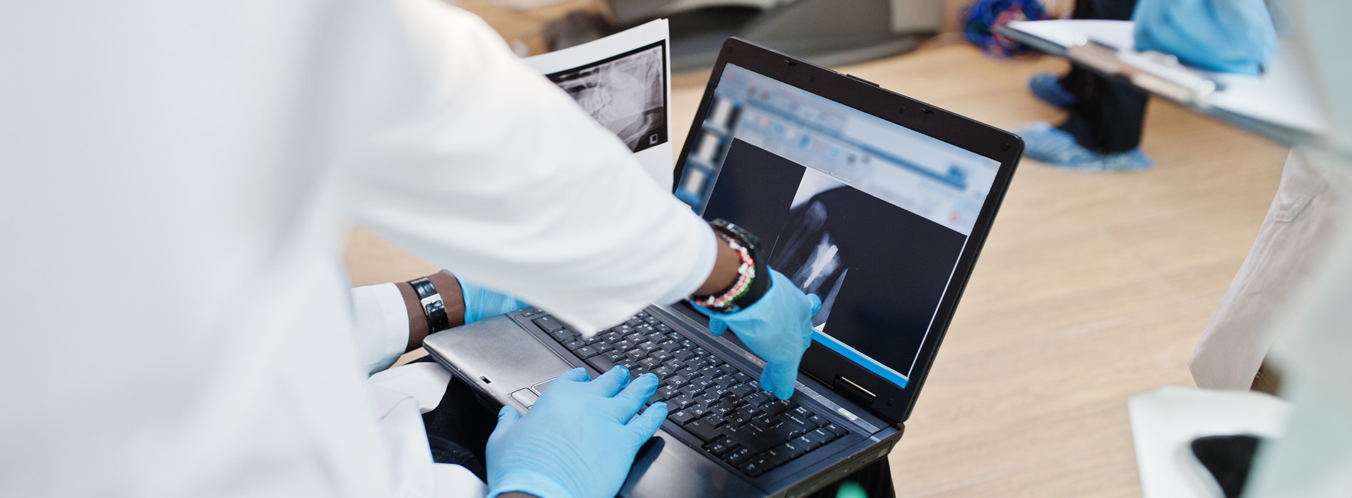 The image features a person working on a laptop with multiple screens displaying various information, set against a backdrop of another person operating a computer workstation with additional laptops and monitors, suggesting a technology-focused environment such as a lab or office setting.