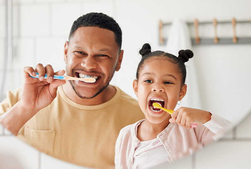 A man and a child are brushing their teeth together in front of a bathroom mirror.