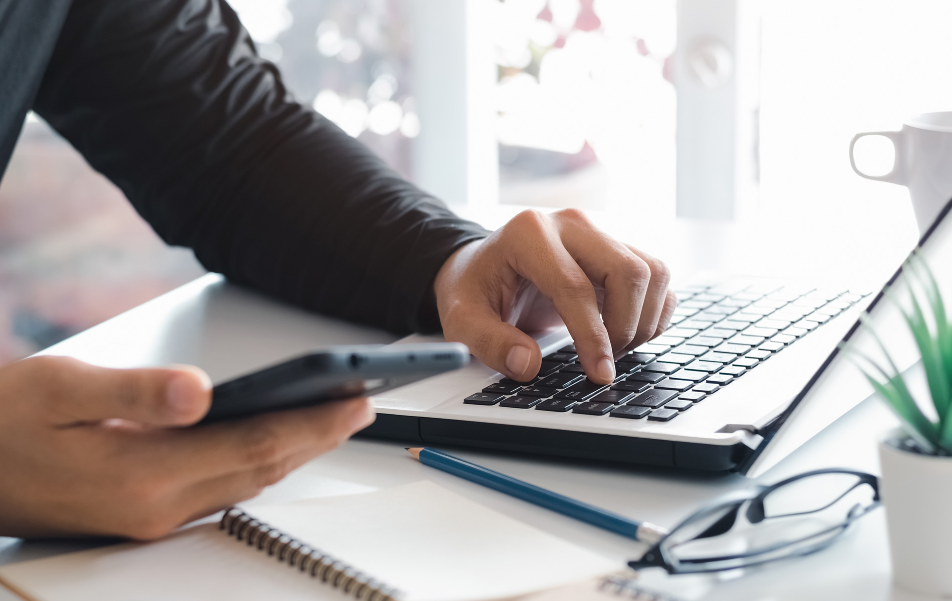 The image shows a person s hands typing on a laptop keyboard with a blurred background featuring a desk and a coffee cup, suggesting a workspace setting.