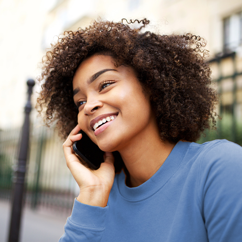 The image shows a smiling woman with curly hair using a cell phone while standing outdoors during daylight hours.