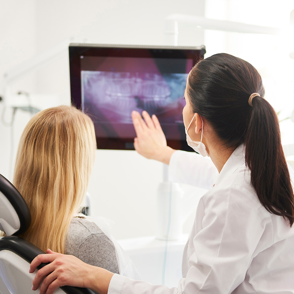 A dental hygienist shows a patient an X-ray of their teeth on a monitor while sitting in a dental chair.