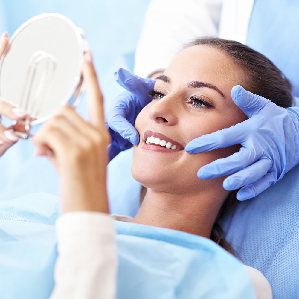 Woman sitting on chair with blue face mask, smiling at camera while looking into large mirror held by person wearing blue gloves.