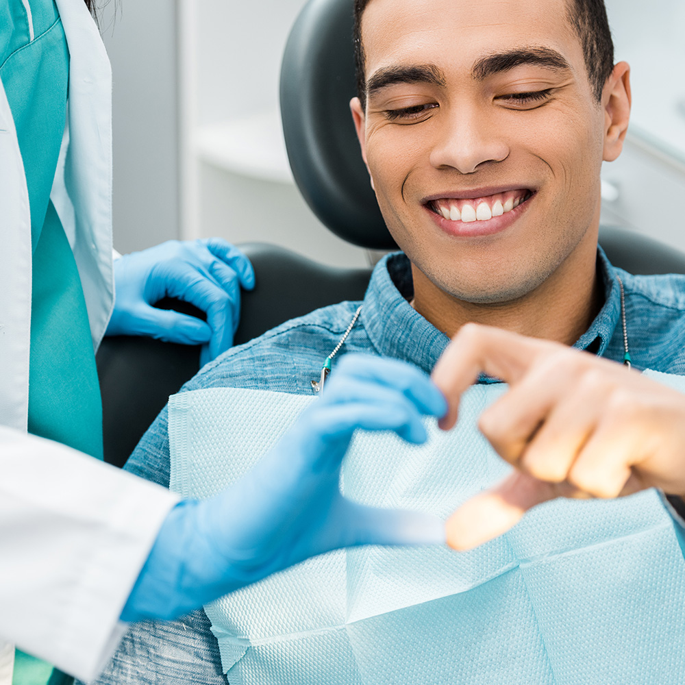 A man seated in a dental chair, smiling broadly at the camera, with his hands forming a heart shape around his chest, while a dental professional attends to him.