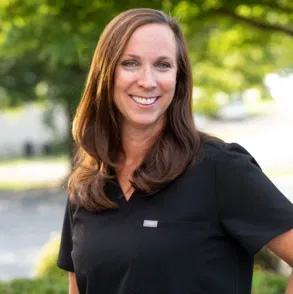 The image shows a woman wearing a black polo shirt with a name tag, smiling at the camera, standing outdoors during daylight.