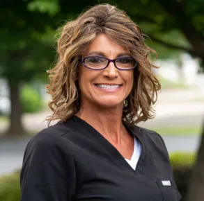 The image shows a woman wearing glasses, with her hair styled in blonde highlights, smiling at the camera. She has a professional appearance, dressed in a white shirt and black pants, suggesting she might be a nurse or healthcare worker.