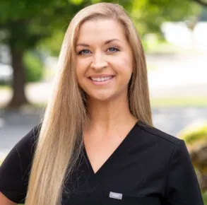 The image shows a woman wearing a black shirt and a white name tag, standing outdoors with trees in the background.