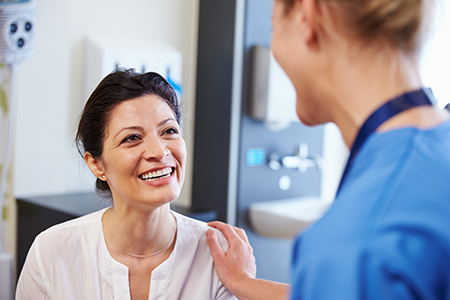 The image shows two individuals, likely in a healthcare setting, with one person seated and smiling at the camera while another stands behind them, possibly a medical professional, engaging in conversation.