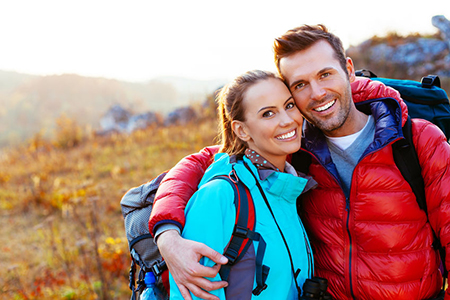 A man and woman are posing outdoors, smiling at the camera, with the man wearing a backpack, suggesting they might be on a hike or outdoor adventure.