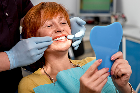A woman sitting in a dental chair with a tooth model held up to her face, smiling at the camera while being attended to by a dental professional.