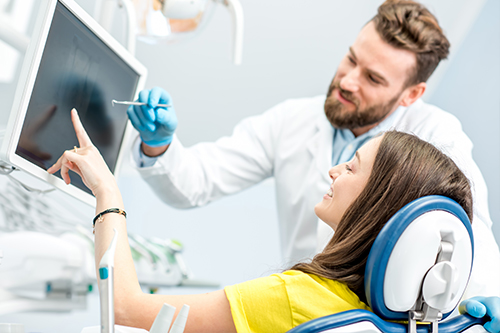 A man in a lab coat assisting a woman seated in a dental chair with a large screen displaying medical images behind them.