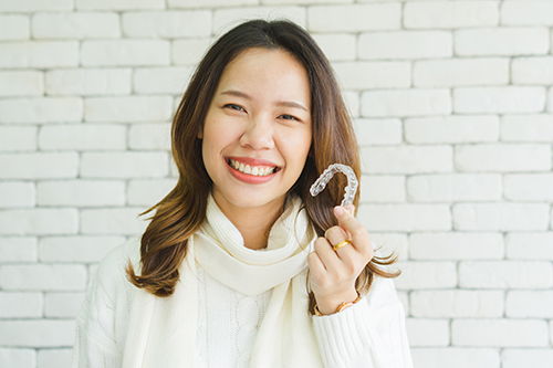 A woman with a smile holds up a hand towards her face while standing against a brick wall.