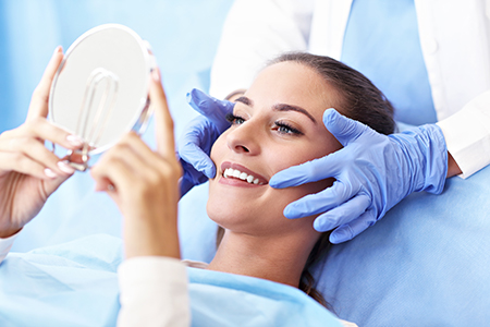 A person sitting on a dental chair with a mirror held up to their face while receiving dental care, with a medical professional performing the procedure.