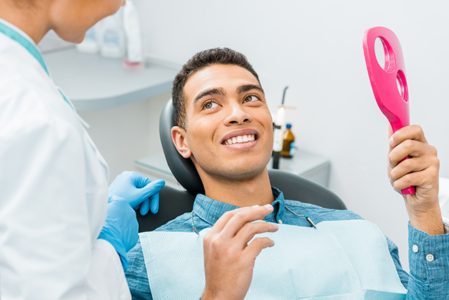 A man sitting in a dental chair, smiling at the camera, with a pink object in his hand, being attended by a dental professional.