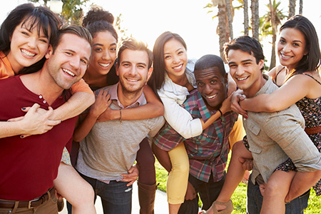 The image shows a group of young adults gathered together, smiling and posing for a photo with their arms around each other, against a sunny outdoor backdrop.