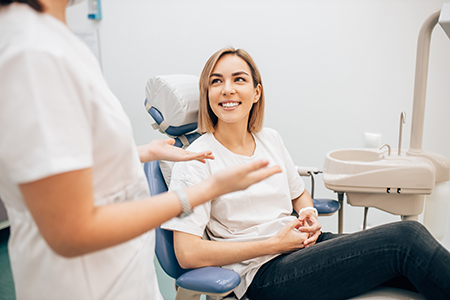 This is a photograph showing a woman seated in a dental chair with her legs crossed, smiling at someone outside the frame, while a dentist s hand is visible holding her arm.