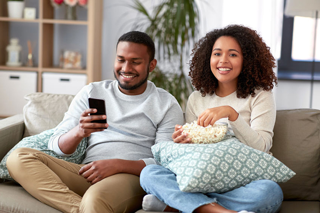 A man and woman sitting on a couch watching TV with snacks, smiling and enjoying each other s company.